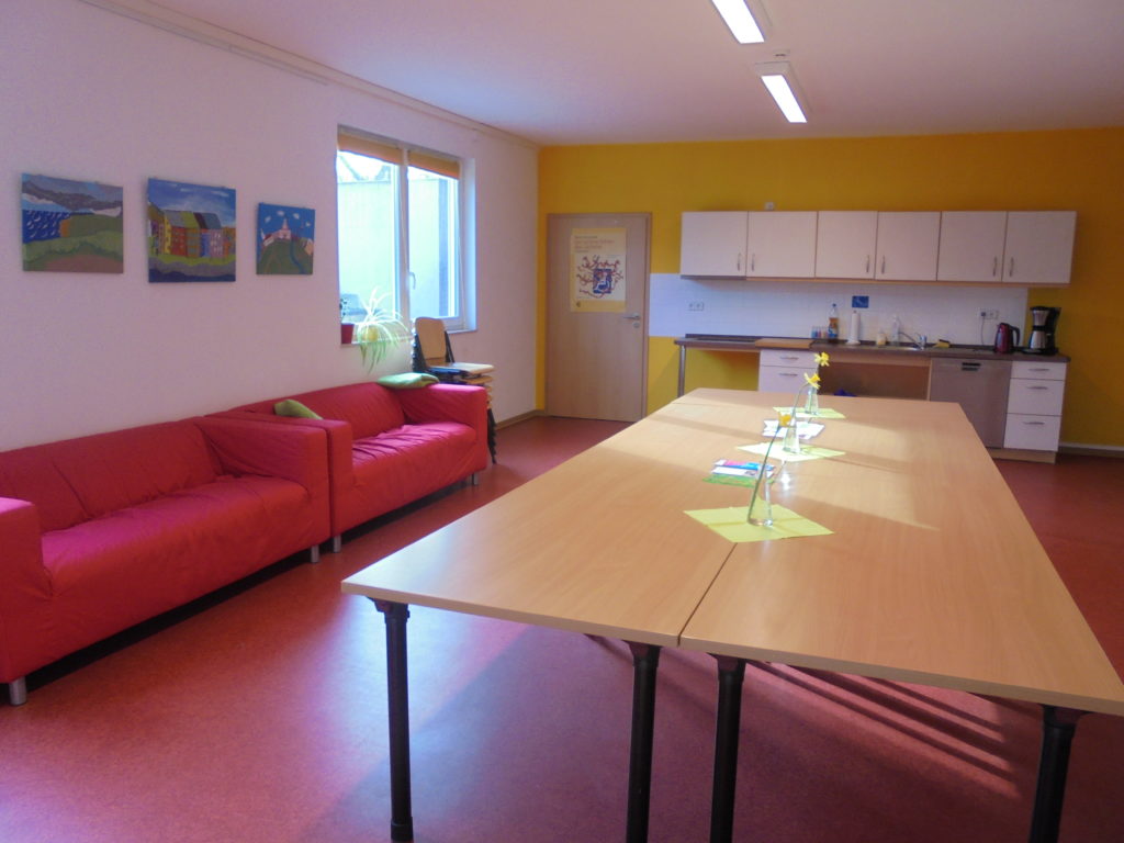 Großer Raum mit langer Tischreihe in der Mitte, einer Küche am Ende des Raumes und zwei Sofas am Rand.