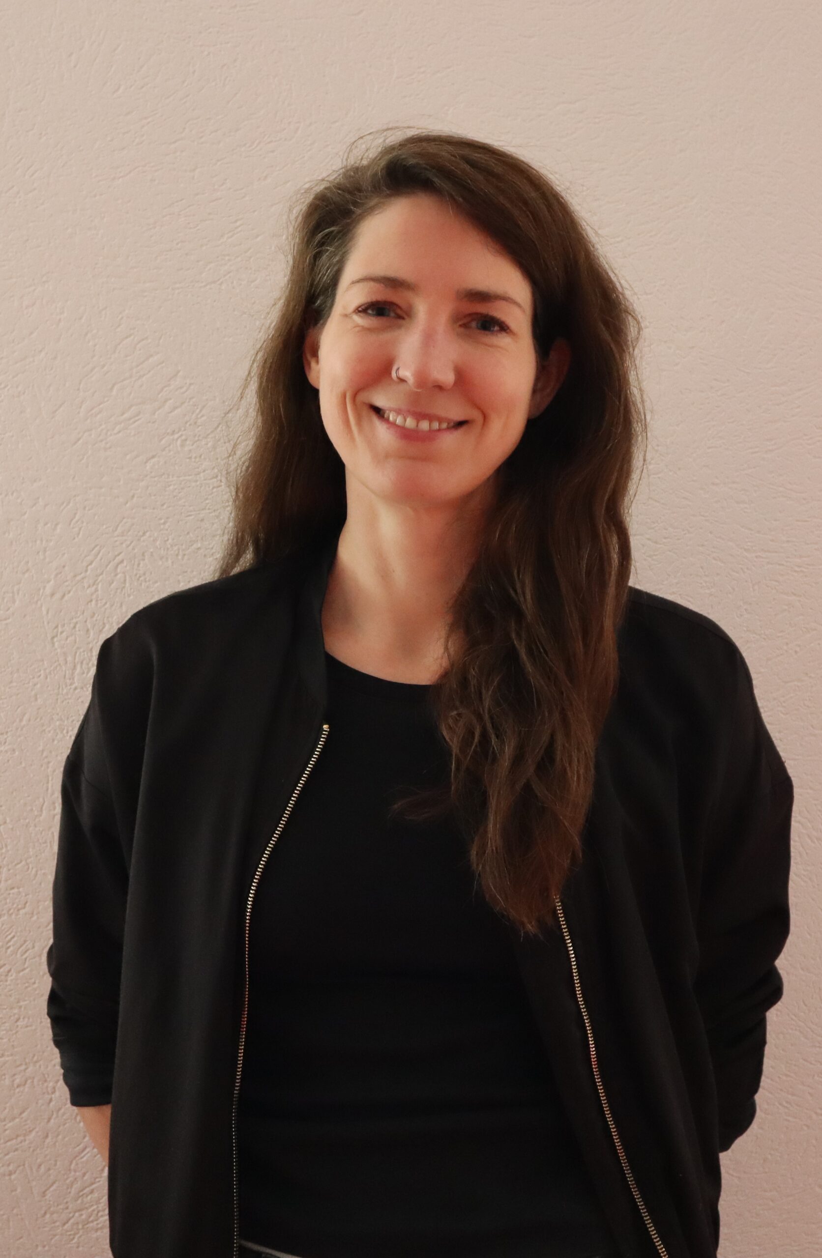 Auf dem Bild ist eine lachende Frau mit langen braunen Haaren zusehen. Sie hat einen Nasenring auf der rechten Seite und trägt eine schwarze Jacke.
