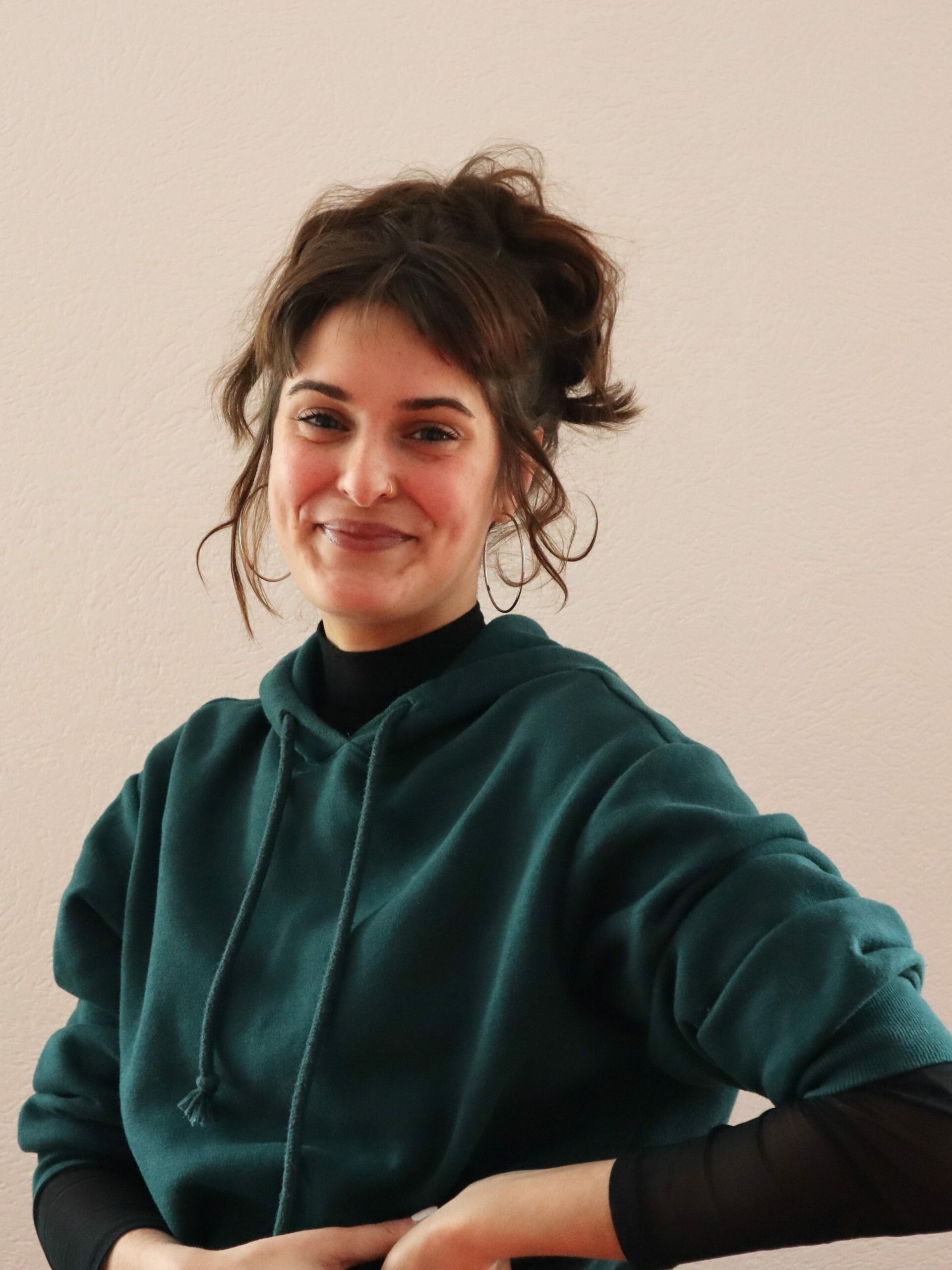 Auf dem Bild ist eine grinsende Frau mit braunen Haaren und Pferdeschwanz zu sehen. Sie hat einen Nasenring auf der linken Seite und trägt einen dunkelgrünen Pullover.