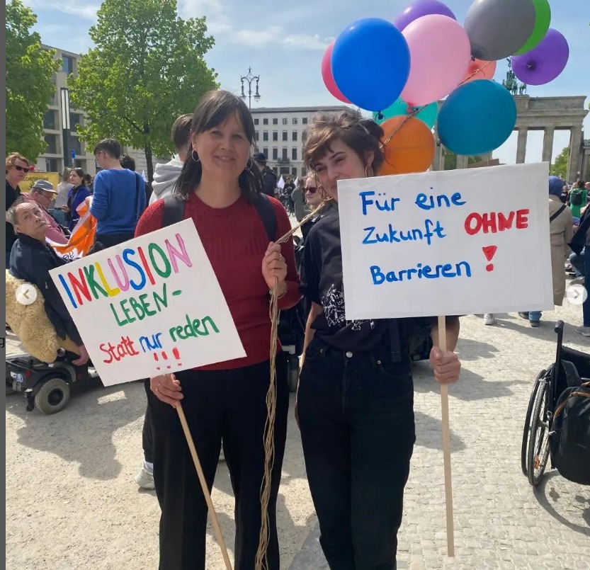 Auf dem Bild zu sehen sidn zwei Frauen. Sie halten jeweils ein Schild in der Hand. Auf dem einen steht: Inklusion leben statt nur reden. Auf dem anderen steht: Für eine Zukunft ohne Barrieren